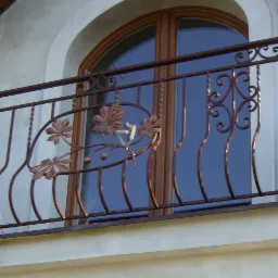 Kuta balustrada balkonowa z motywem liści kasztanowca, brązowa, na tle okna z łukiem i fragmentu dachu.